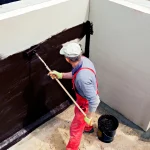A contractor wearing protective gear applying waterproofing material to the exterior wall of a building foundation