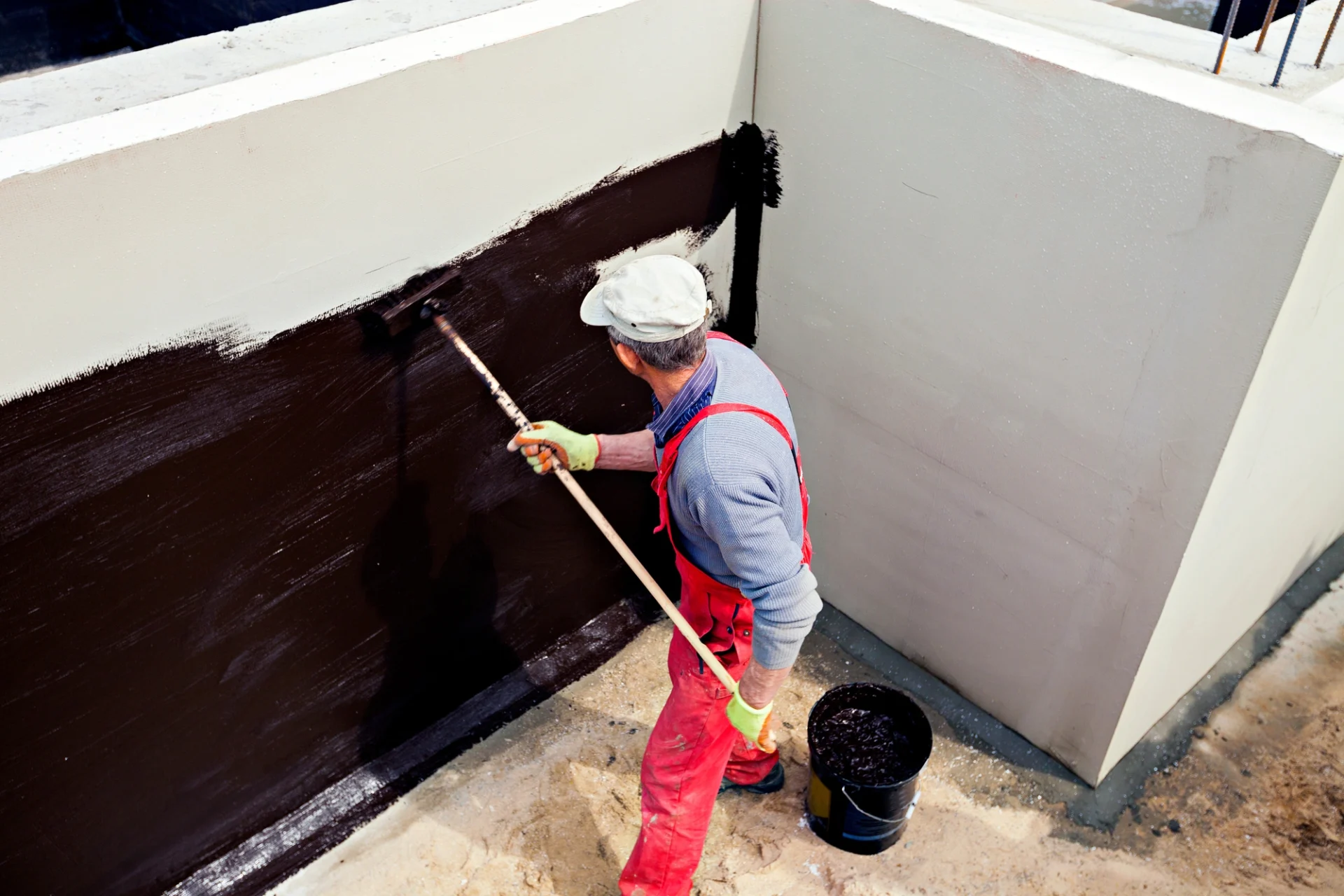 A contractor wearing protective gear applying waterproofing material to the exterior wall of a building foundation
