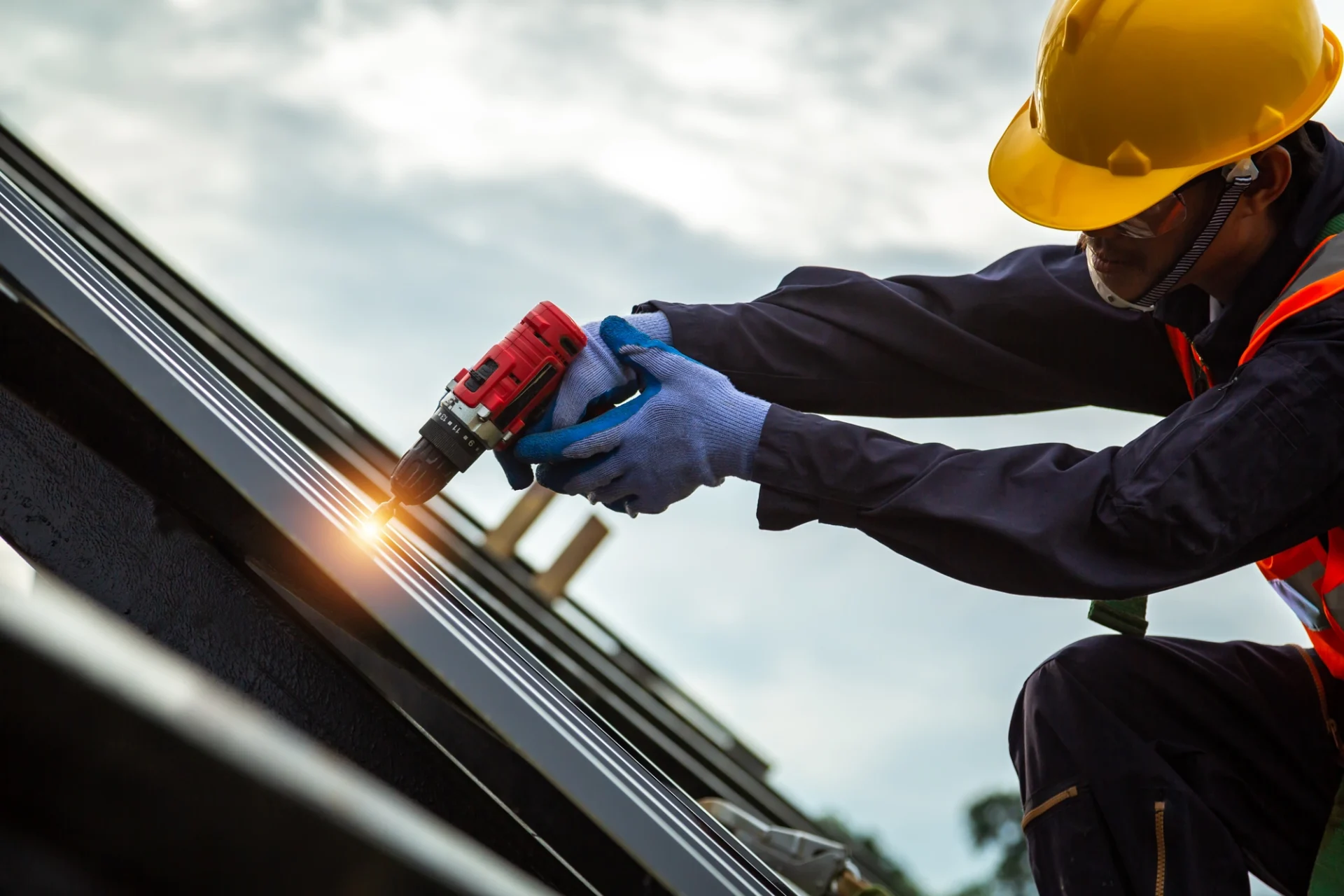 A roofing contractor wearing protective gear using an electric drill on a metal roof