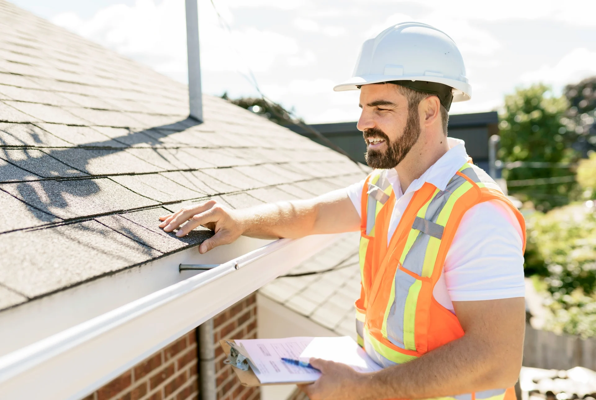 A roofing contractor with a clipboard inspecting roof shingles with a smile