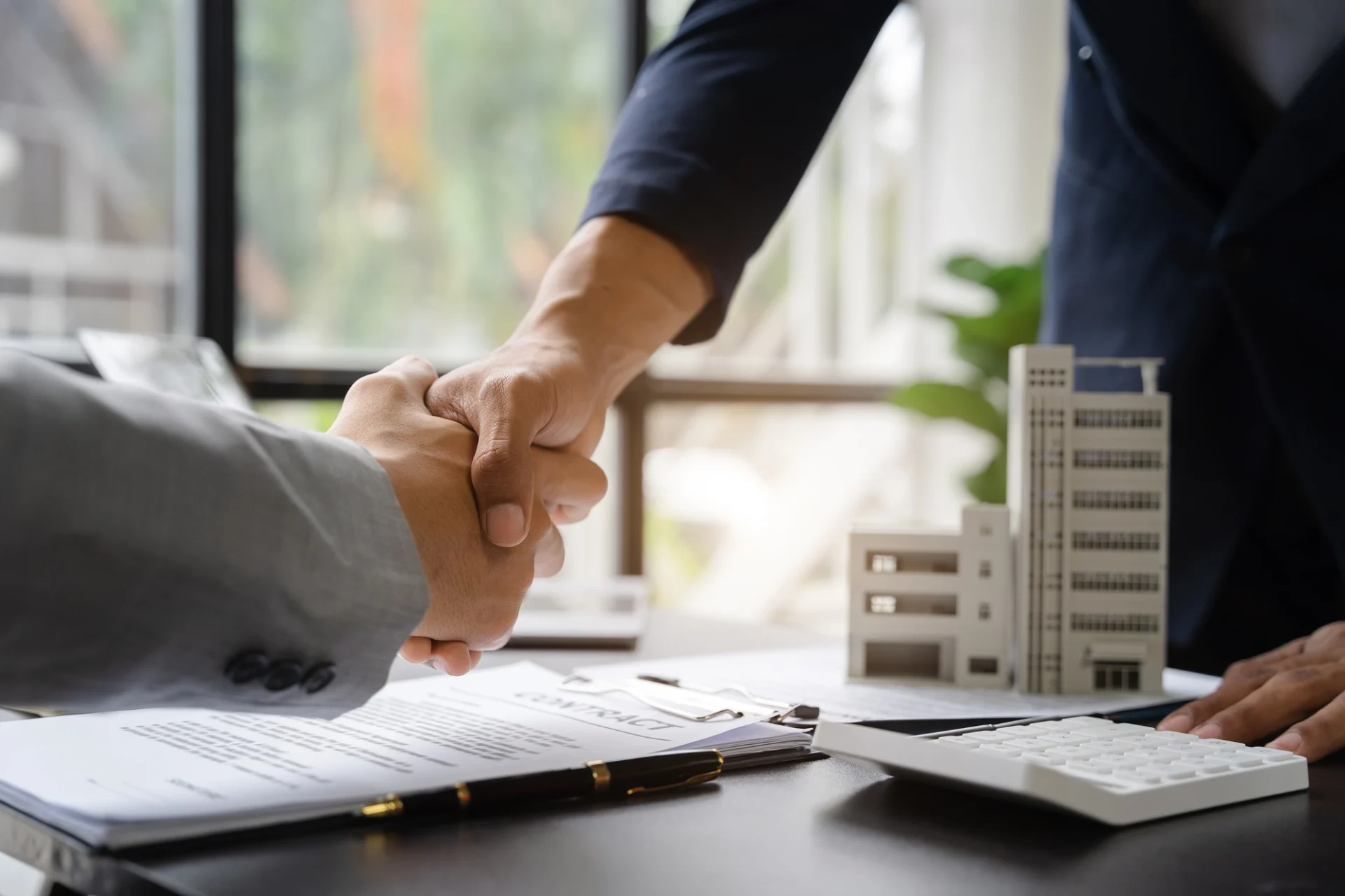 A close-up view of two people shaking hands over a desk with a building model and a contract